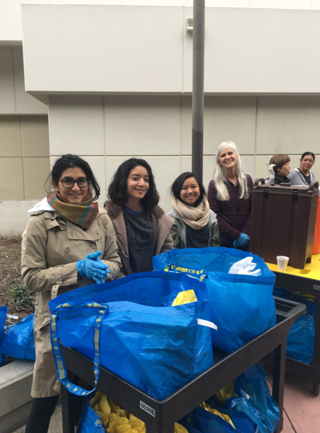 A group of women are standing next to a cart filled with blue bags.