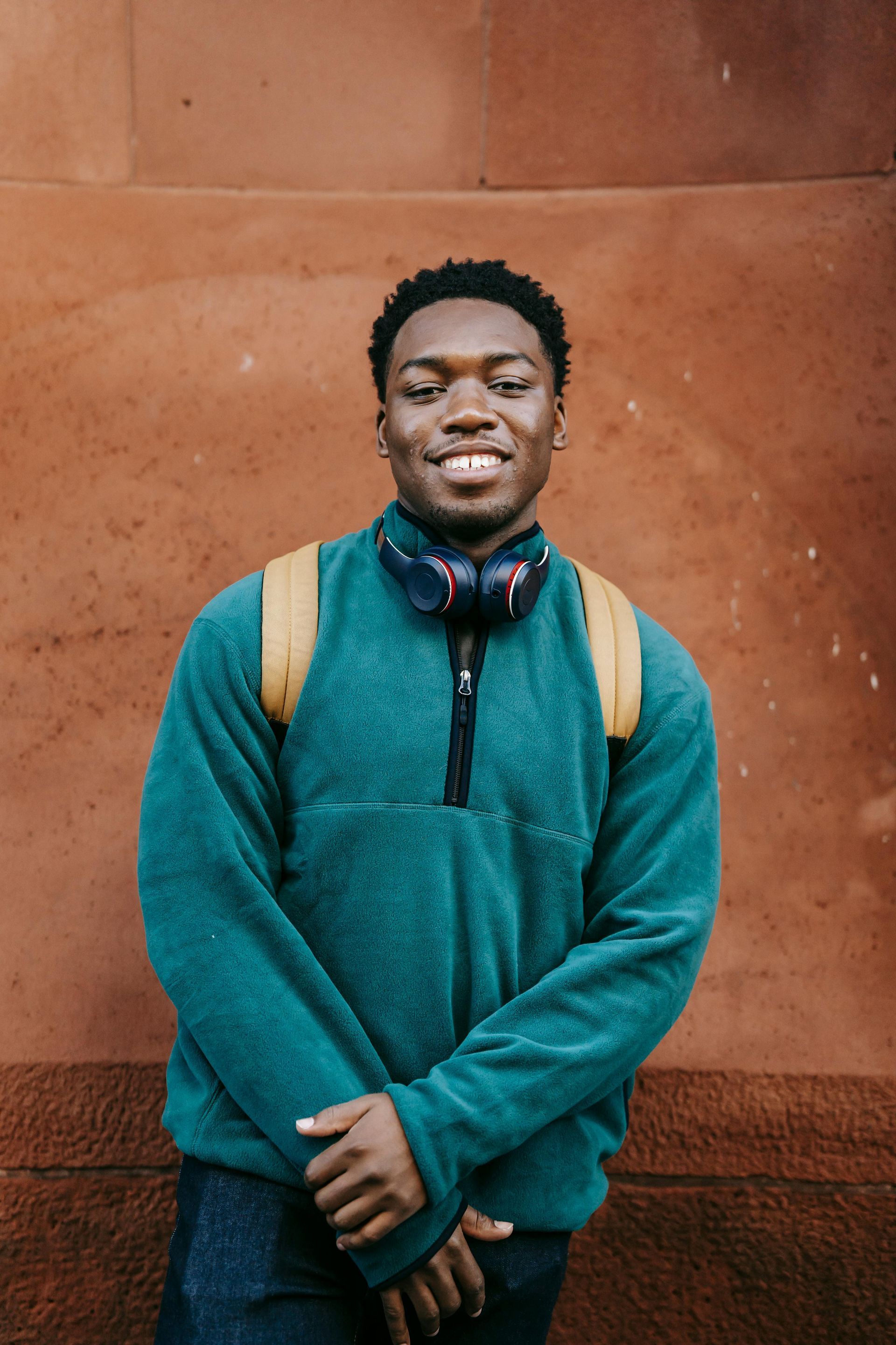 A young man wearing a green sweater and headphones is standing in front of a red wall.