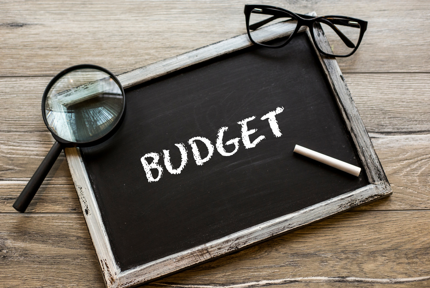 The word budget written on a blackboard with a piece of chalk, glasses, and magnifier lens on a wooden background.