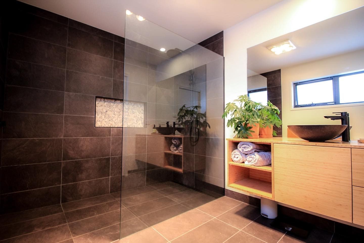 Modern bathroom with dark tiled shower, glass partition, light wood vanity, vessel sink, and potted plant on the counter.