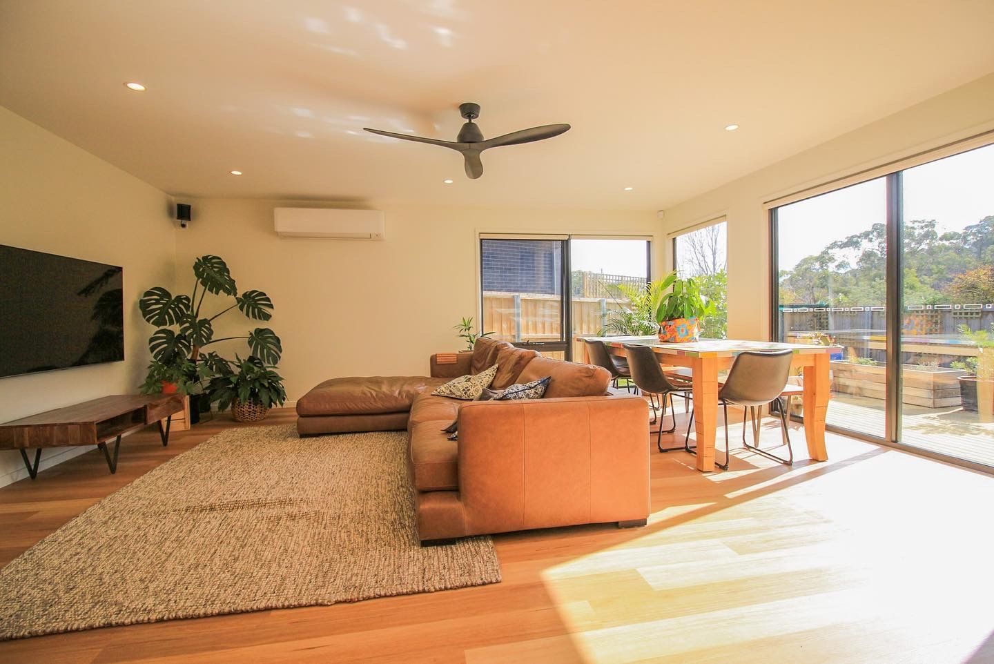 A modern living room with a brown sectional, wooden dining set, large windows, and a television on a wall.