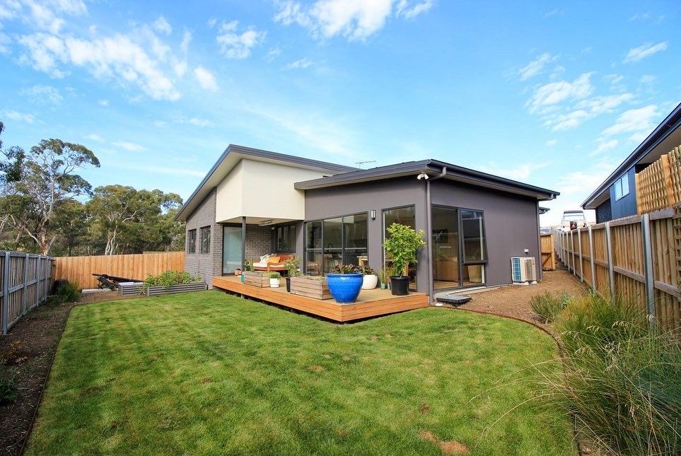 Modern grey house with a wooden deck and green lawn in a backyard under a bright blue sky.