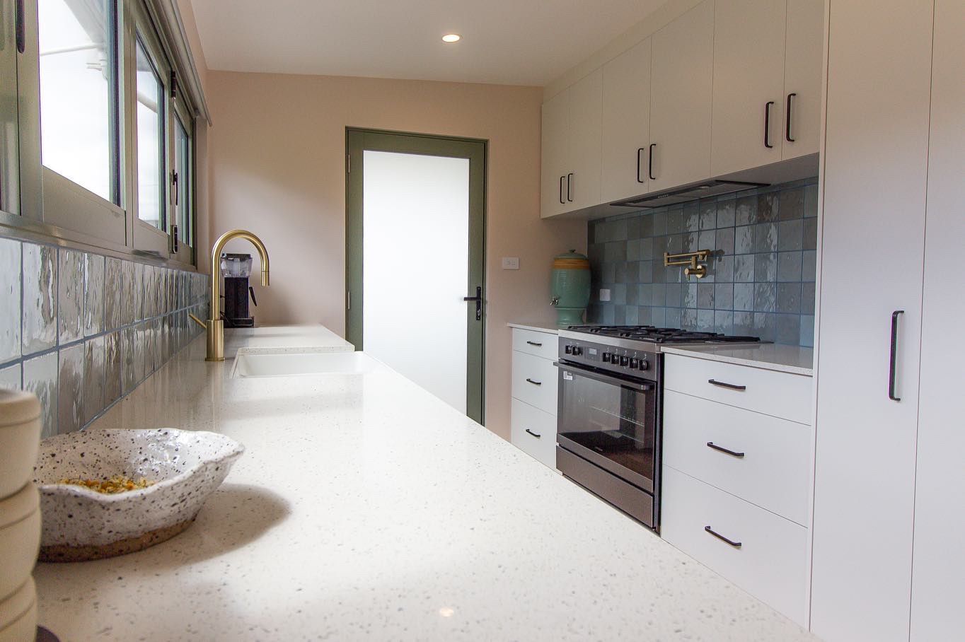 A modern kitchen with white cabinetry, light stone countertops, a gold faucet, and blue-gray subway tile backsplash.