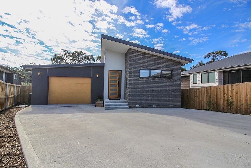 A modern, single-story house with a slanted roof, dark textured siding, a wooden garage door, and a concrete driveway.