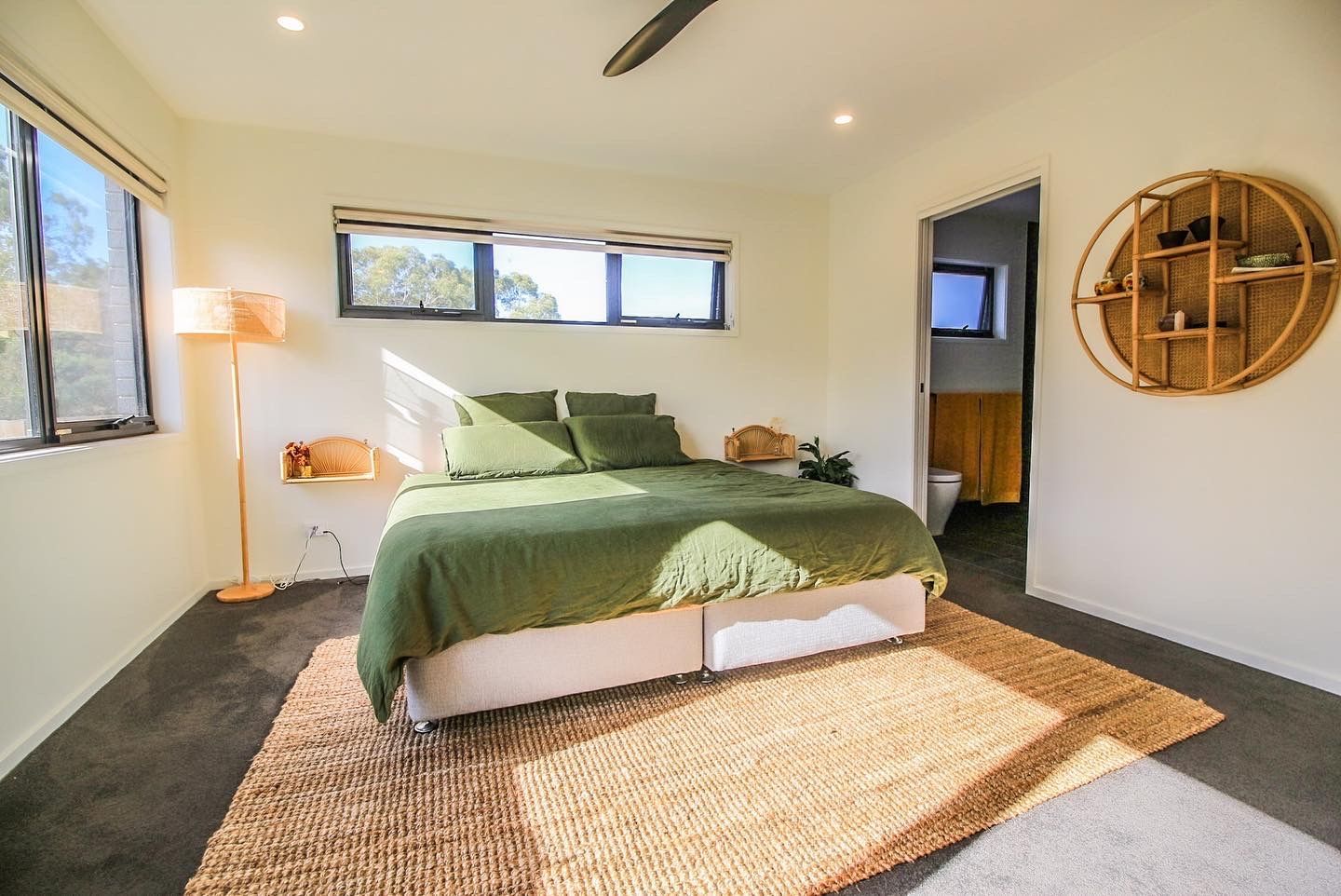 A bedroom with a green bed, a round wooden wall shelf, a jute rug, and a floor lamp against a bright, neutral wall.
