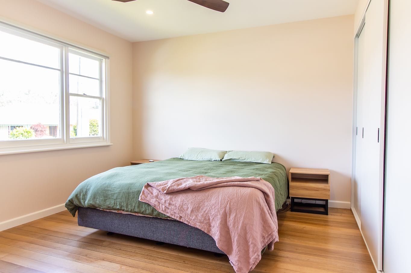 A minimalist bedroom with light walls, a bed featuring green bedding and a pink throw, and two wooden nightstands.