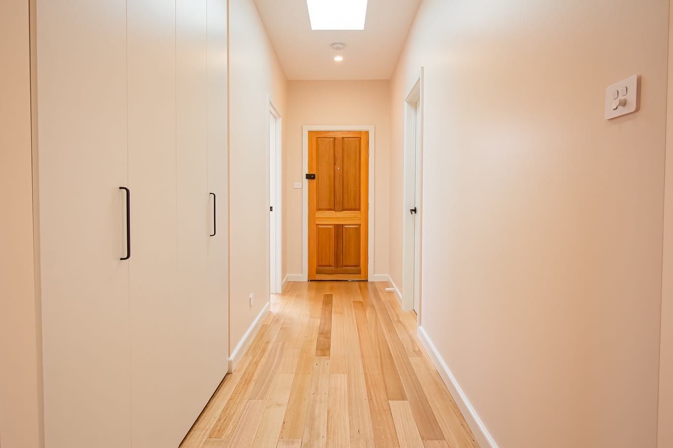 A bright hallway with light wood flooring, white walls, built-in closets on the left, and a wooden door at the end.