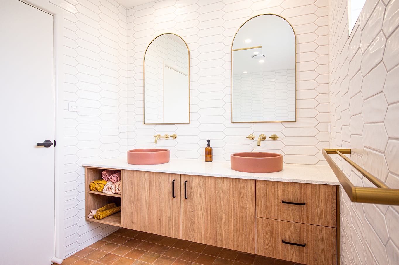 A modern bathroom vanity with wood cabinets, two pink round sinks, gold faucets, and arched mirrors on white tiled walls.