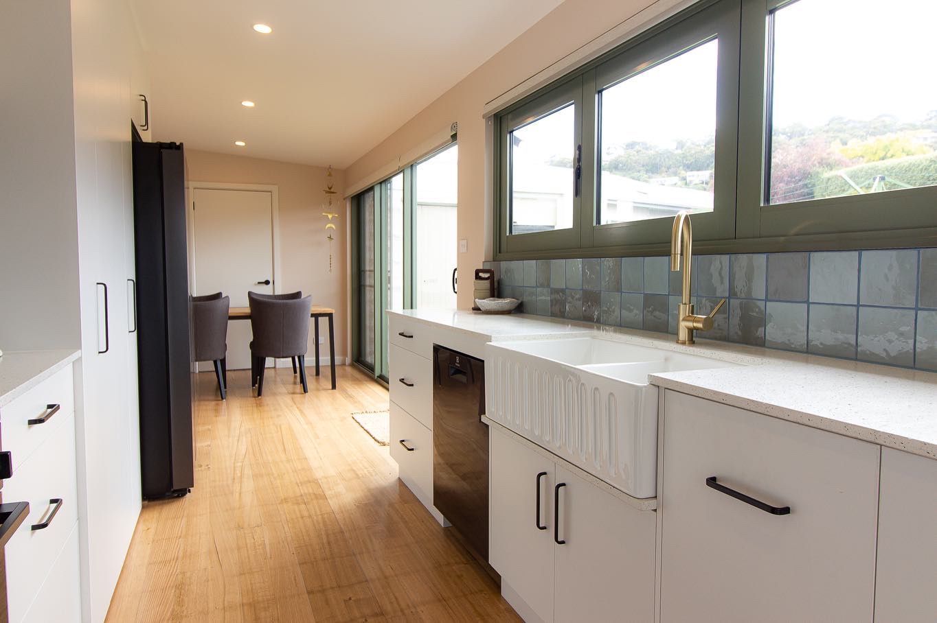 A bright, modern kitchen featuring white cabinets, a farmhouse sink, gold faucet, blue tile backsplash, and wood flooring.
