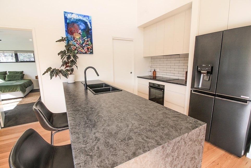 Modern kitchen with a dark stone island, black stools, black refrigerator, and a glimpse of a bedroom in the background.