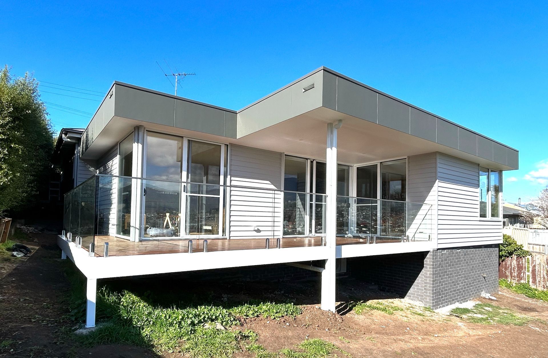 Modern elevated house with a white deck, large glass windows, and grey siding against a clear blue sky.