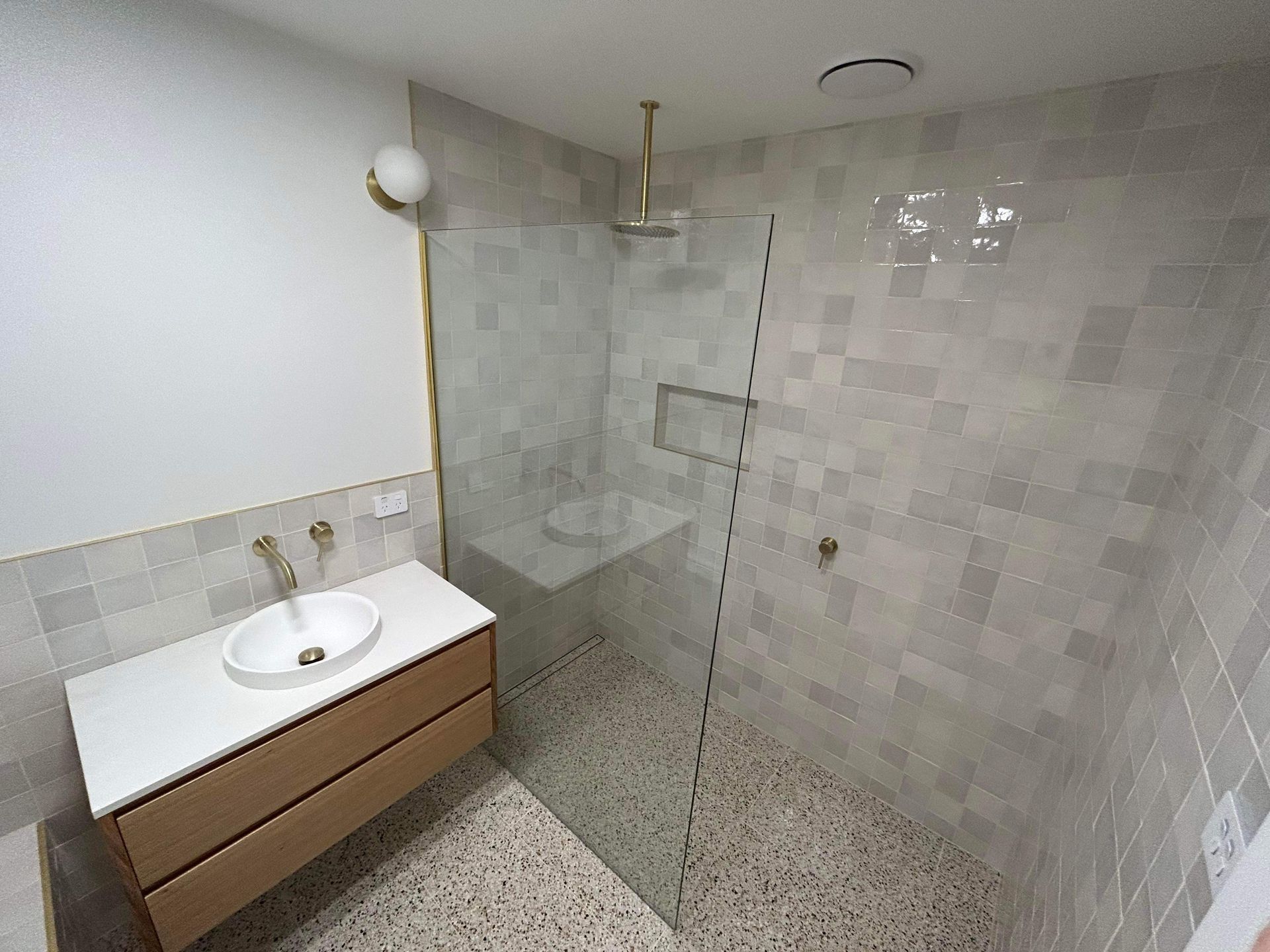 Modern bathroom with a wooden vanity, white countertop, sink, and a walk-in shower with tiled walls and terrazzo floors.