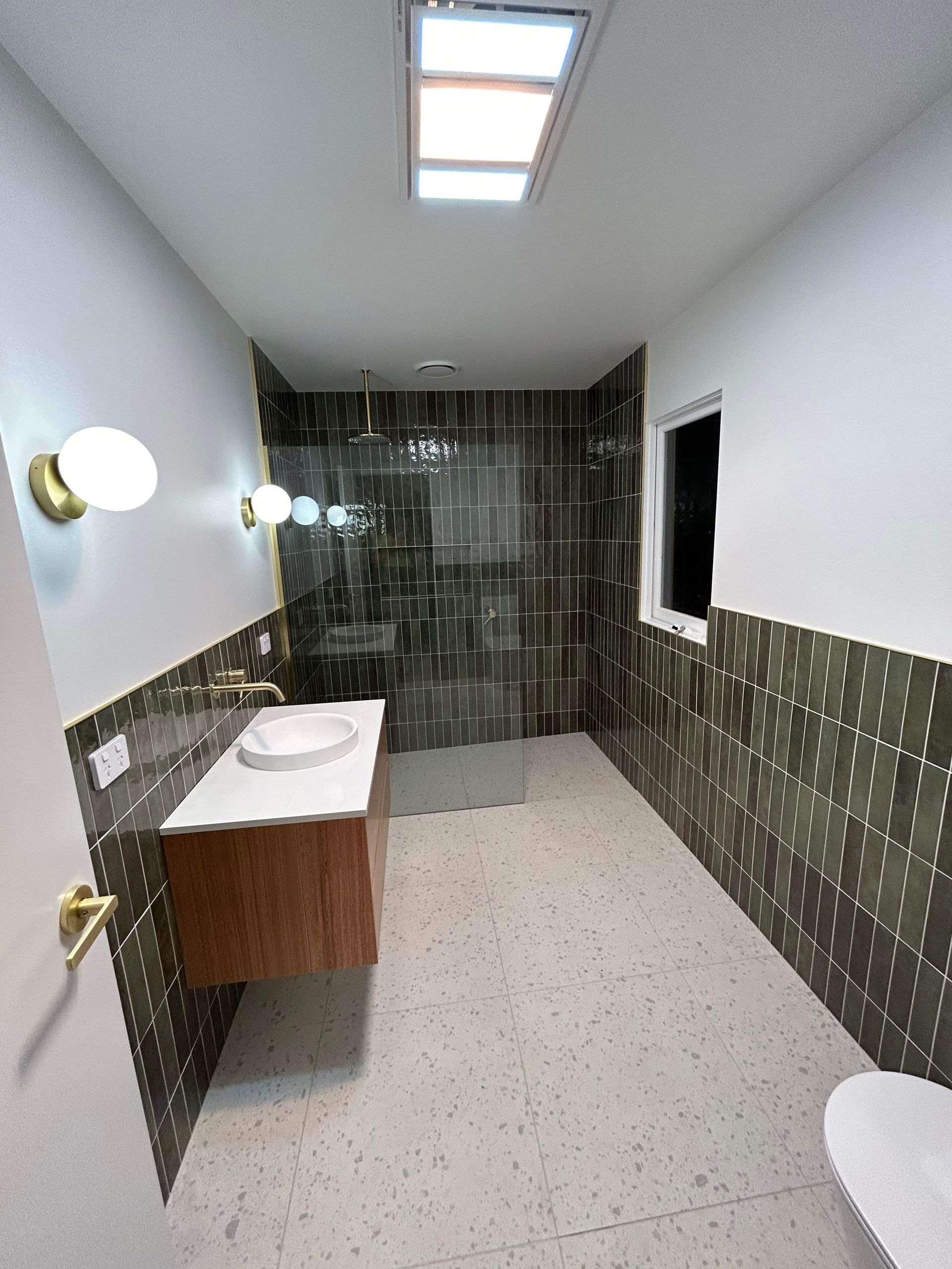 A modern bathroom with dark green tiled walls, light terrazzo flooring, a wooden vanity, and a walk-in glass shower.