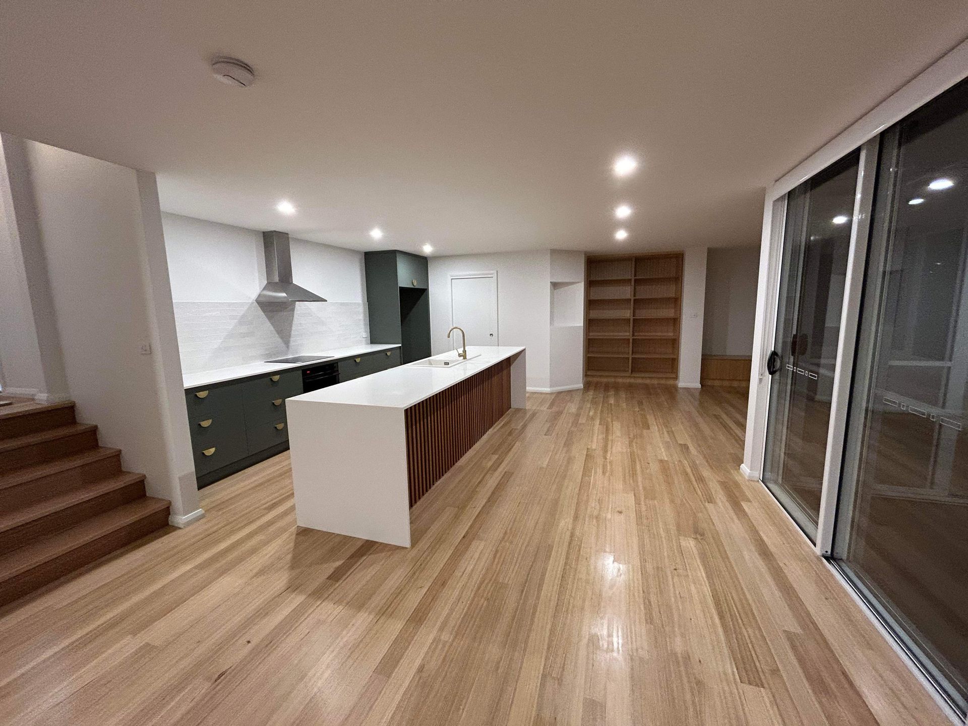 A modern, open-plan kitchen with dark green cabinetry, a white island, wooden flooring, and a large sliding glass door.