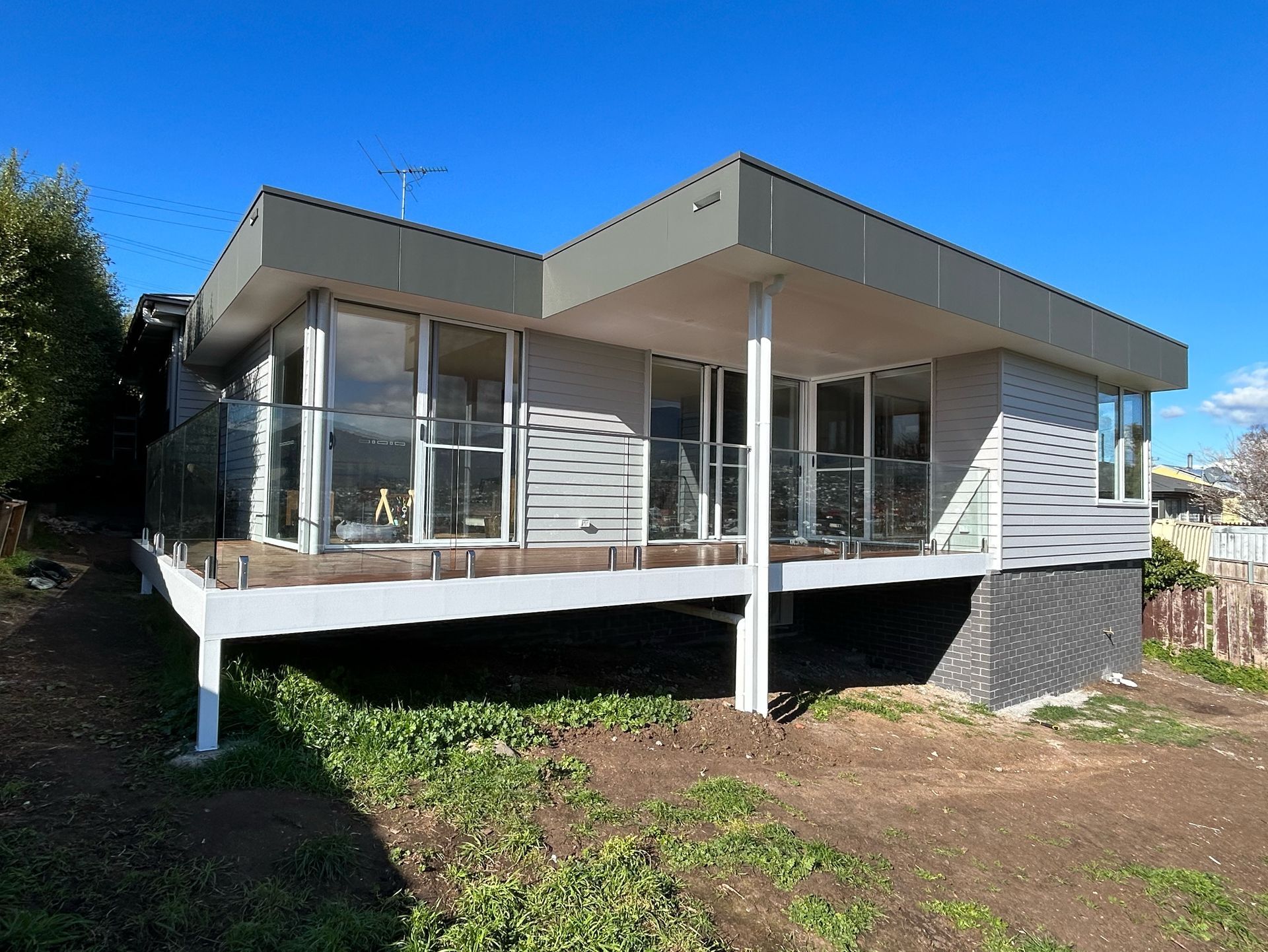 A modern, elevated house with light-colored siding, a metal roof, and a glass-railed deck on a sunny day.