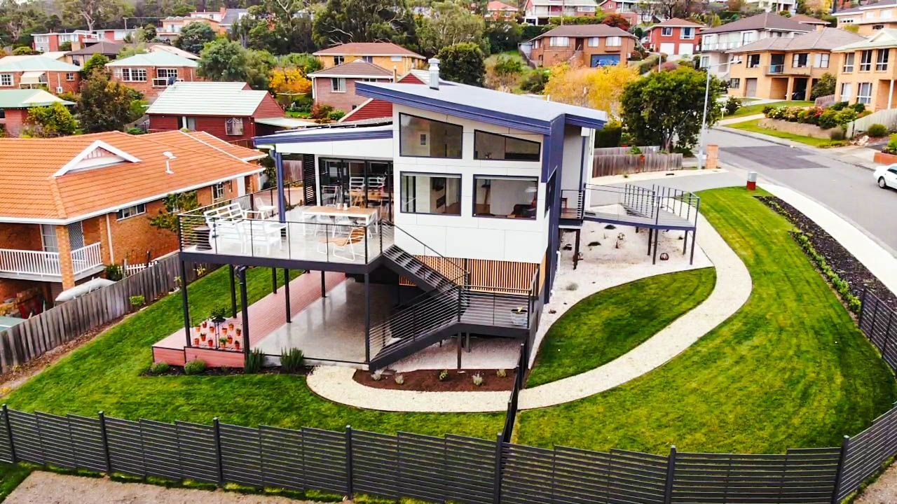 A modern, two-story house with a blue roof, raised deck, and sweeping lawn in a residential neighborhood.