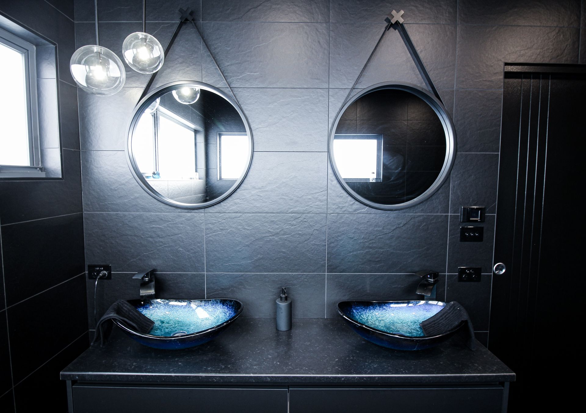 A modern bathroom with a black vanity, two blue glass vessel sinks, and round mirrors hanging on a dark tiled wall.