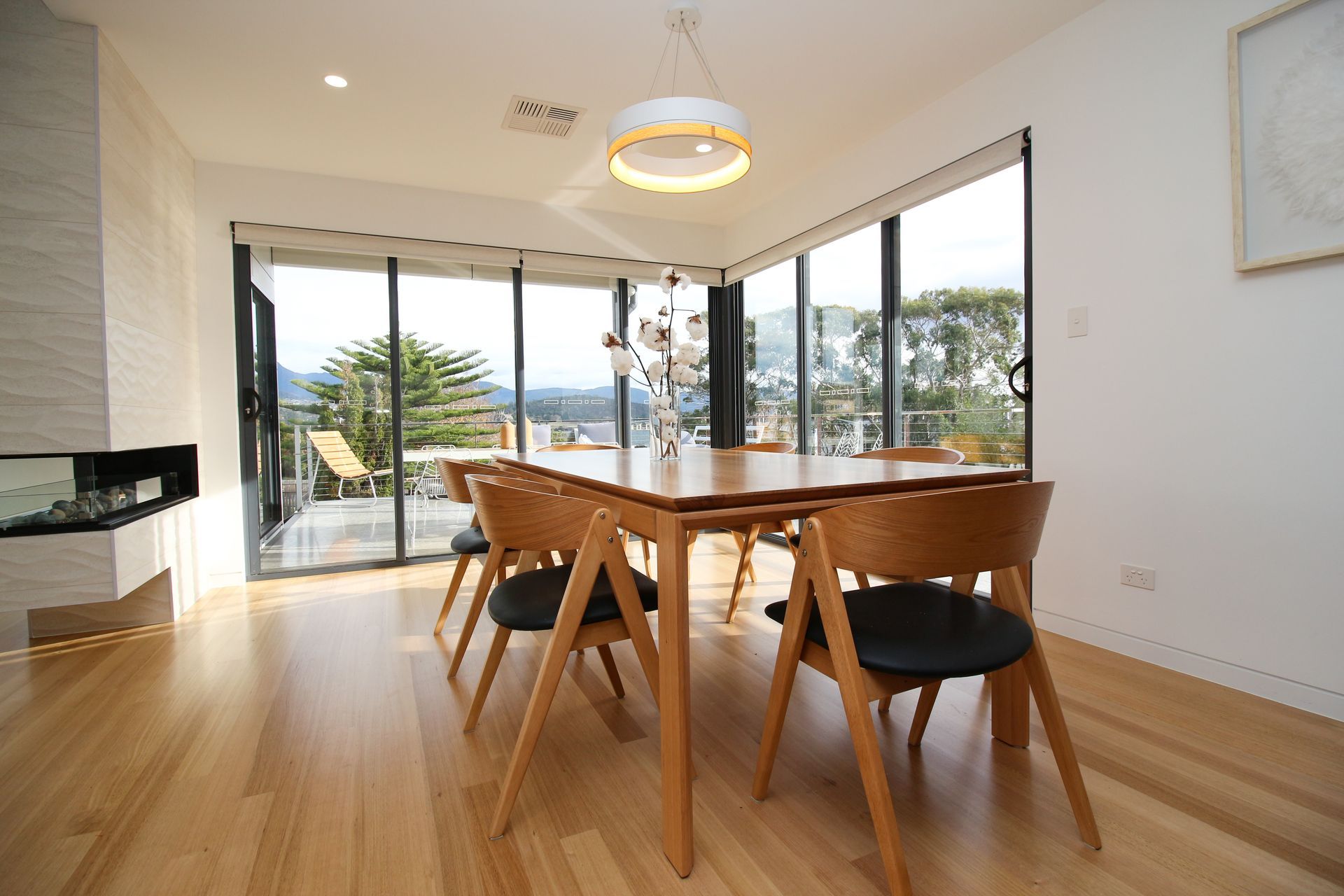 Modern dining area with a wooden table, chairs with black cushions, large windows, and a view of trees and mountains.