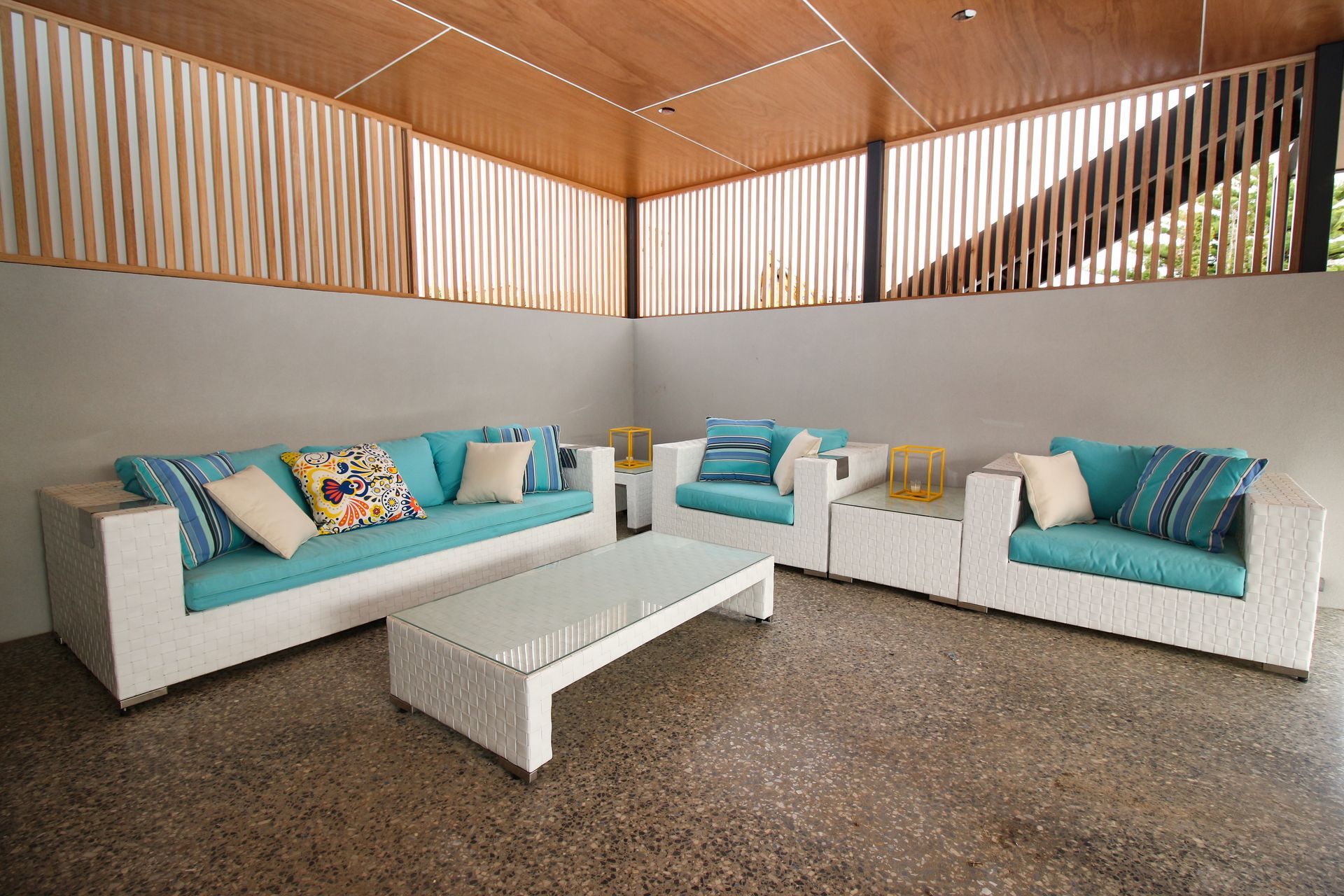 A white wicker lounge set with teal cushions and patterned pillows sits on a stone floor under a wooden-slatted canopy.