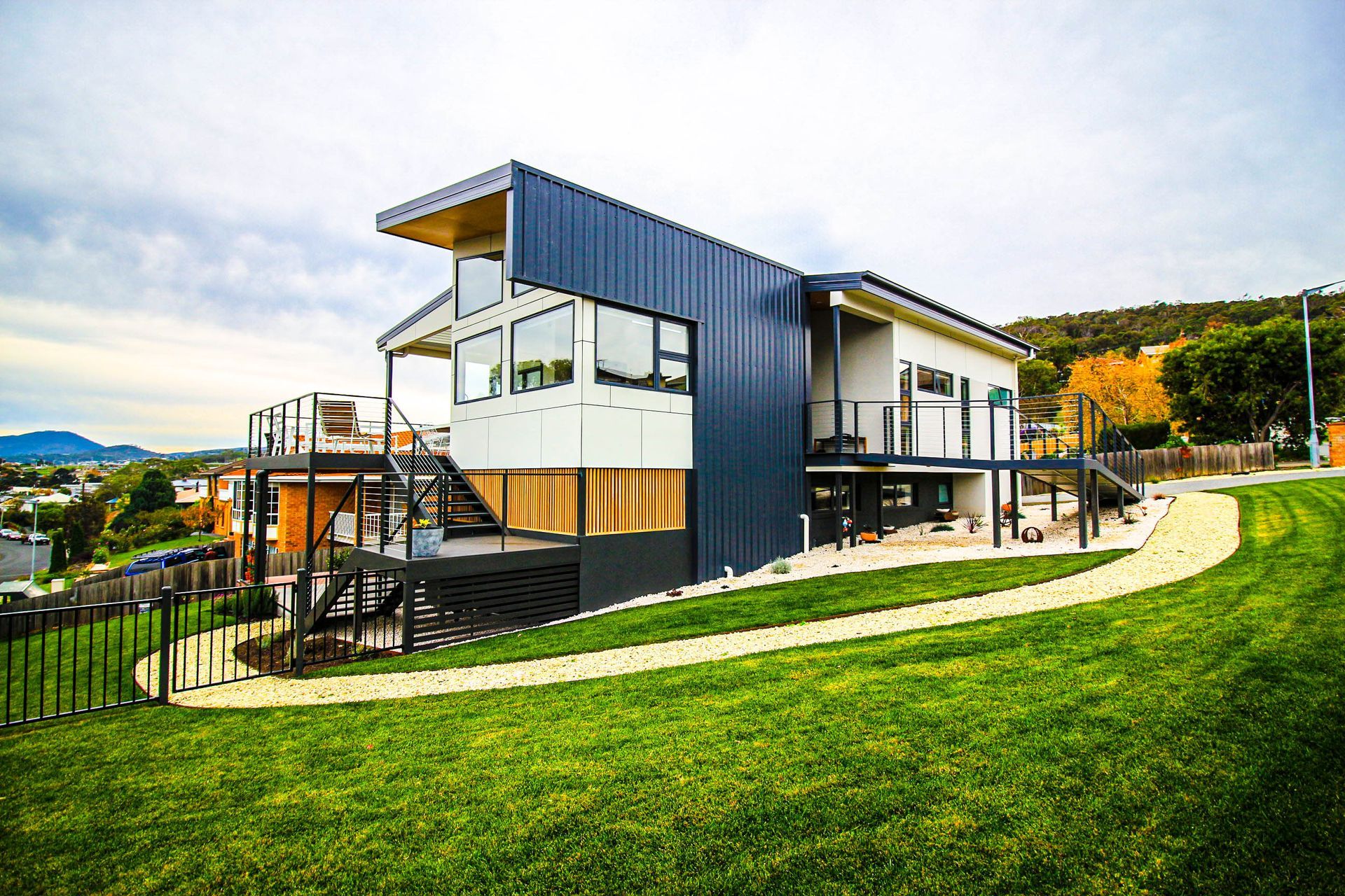 A modern multi-level house with dark metal siding and wood accents, situated on a grassy hill under a cloudy sky.