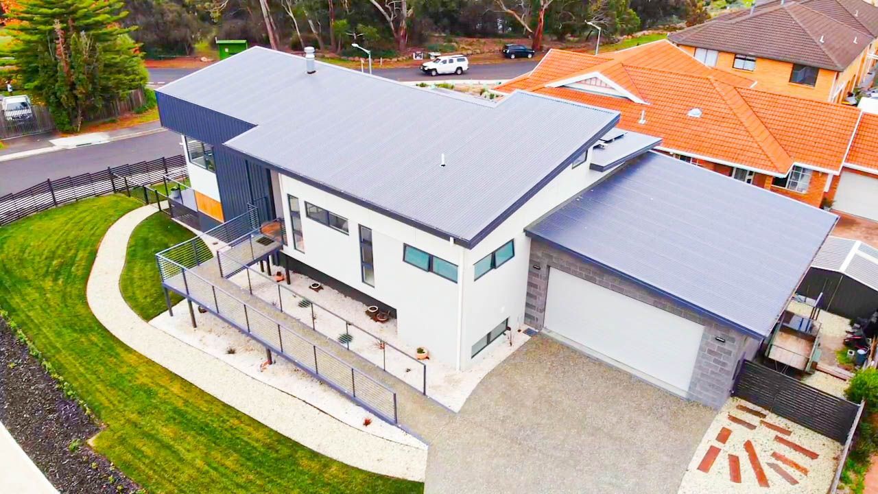 Aerial view of a white, modern two-story house with a grey roof, a garage, and a ramp-style walkway in a suburban area.