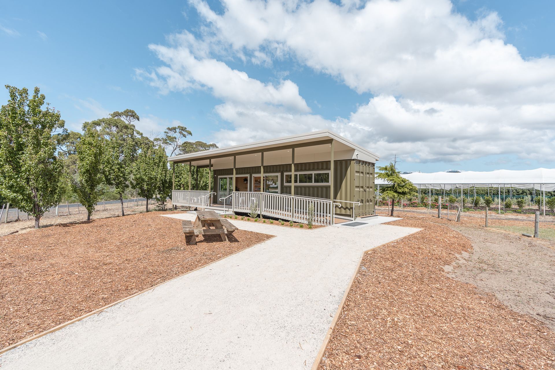 A modern, olive-green cabin sits in a landscaped orchard, accessible via a light-colored gravel path and wooden ramp.