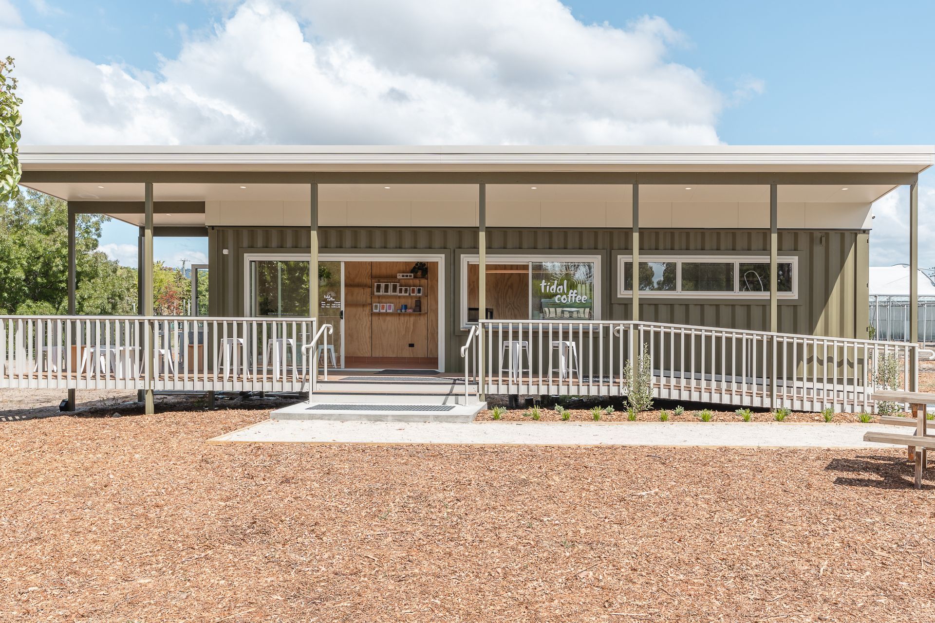 A olive-green modular building with a wide porch and wheelchair-accessible ramp, set in a wood-chipped outdoor area.