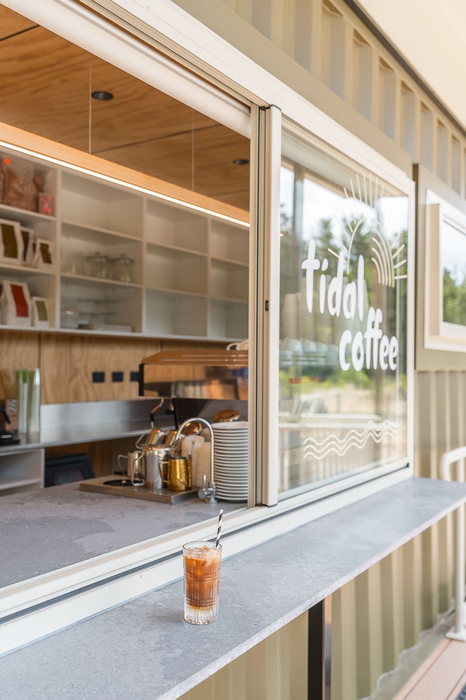 A single iced coffee sits on the service counter of a Tidal Coffee stand, featuring a light-toned wood and metal interior.