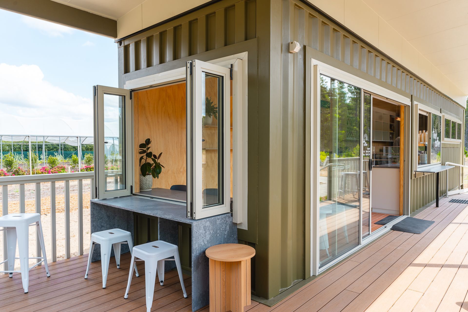 An olive-green shipping container home with an outdoor deck, a folding window serving as a bar, and white stools.
