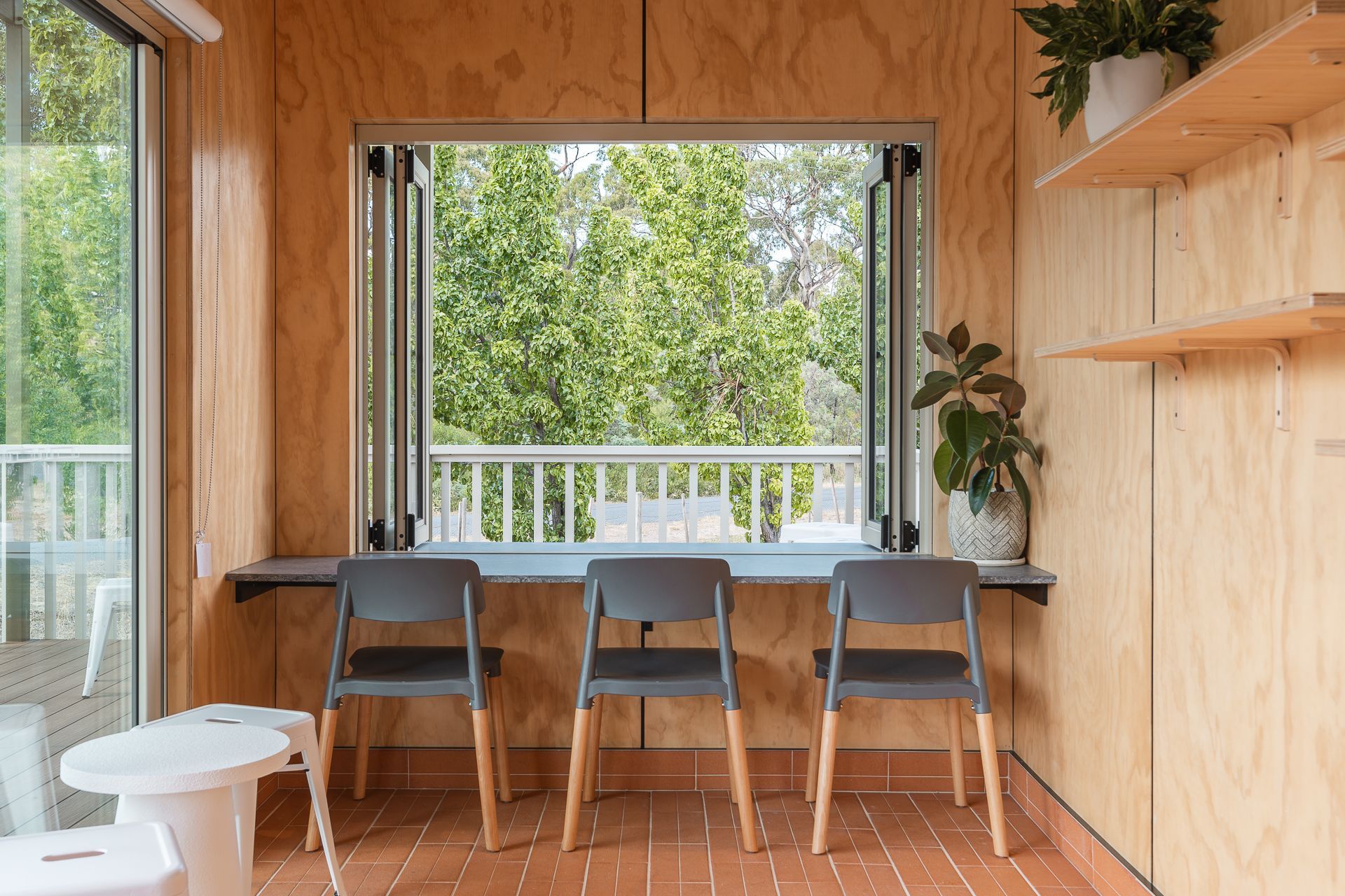 A room with wooden walls, three gray chairs at a window desk, a floor-to-ceiling glass door, and a potted plant.