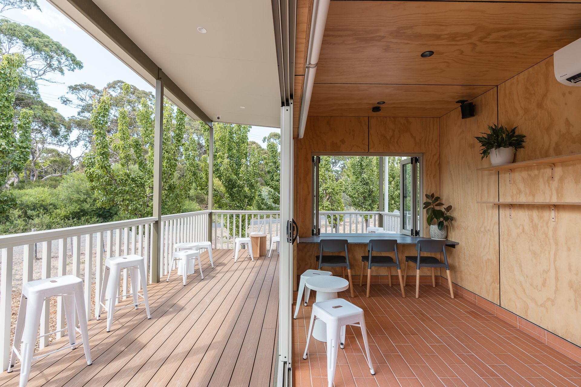 An outdoor wooden deck and covered area with white stools and a built-in counter overlooking a green, tree-filled landscape.