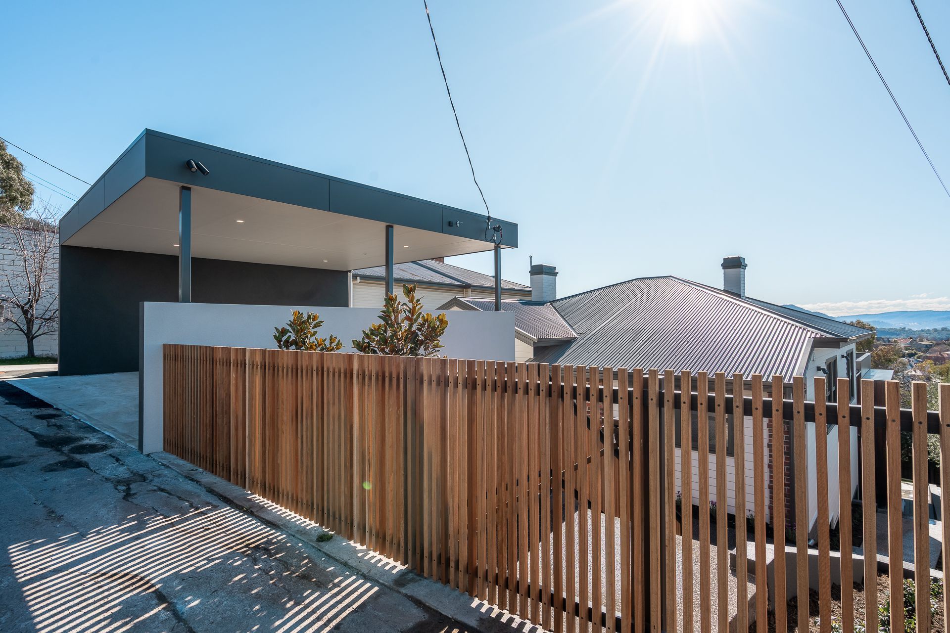 A modern dark-gray carport with an attached wooden slat fence sits in front of a residential house under a bright sun.