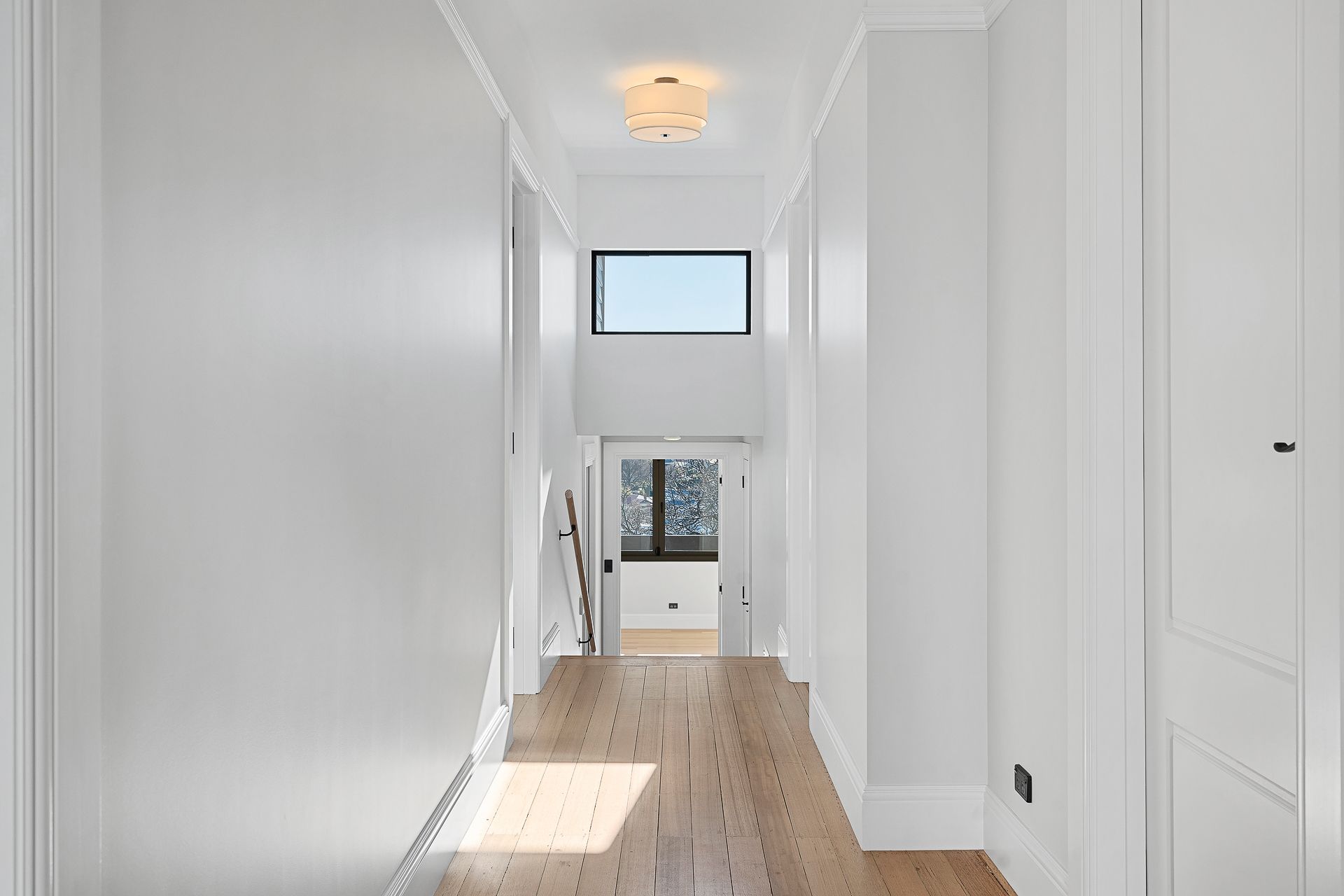A bright hallway with white walls and wood floors, featuring a light fixture, a small upper window, and a glass-paneled door.
