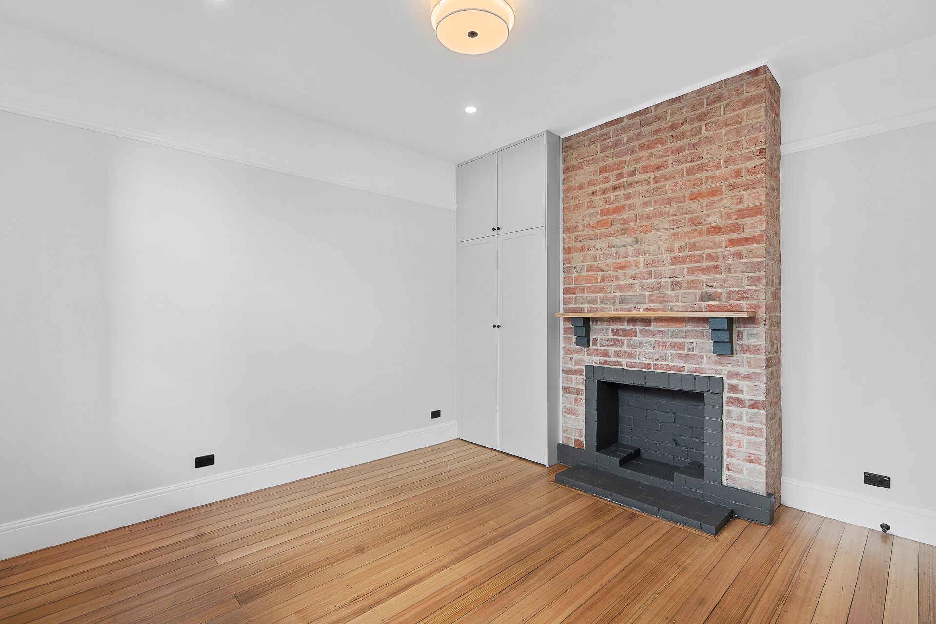 A bright, empty room with light-colored walls, hardwood floors, a exposed brick fireplace, and white floor-to-ceiling cabinets.