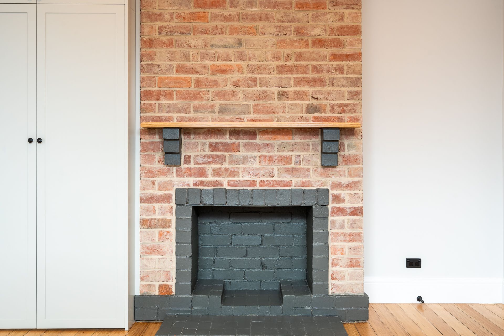 A brick fireplace with a grey painted interior and base, featuring a simple wooden mantel shelf against a white wall.