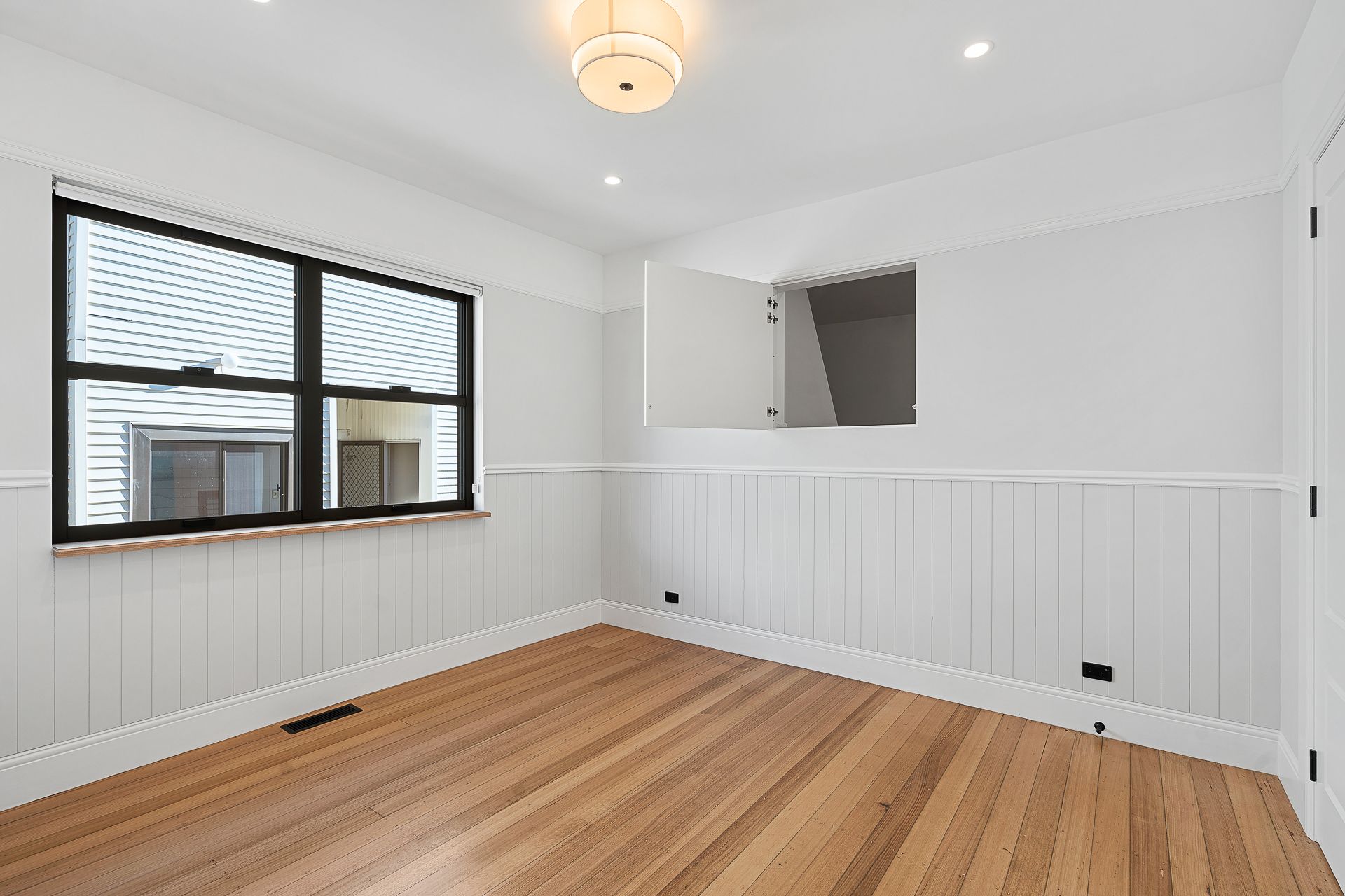 Empty room with white wainscoting, light wood floors, a black-framed window, and a small open wall hatch.
