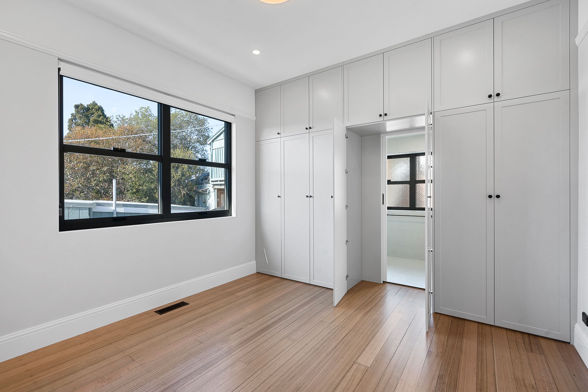 Empty room with light wood floors, gray walls, a window, and built-in floor-to-ceiling white storage cabinets.