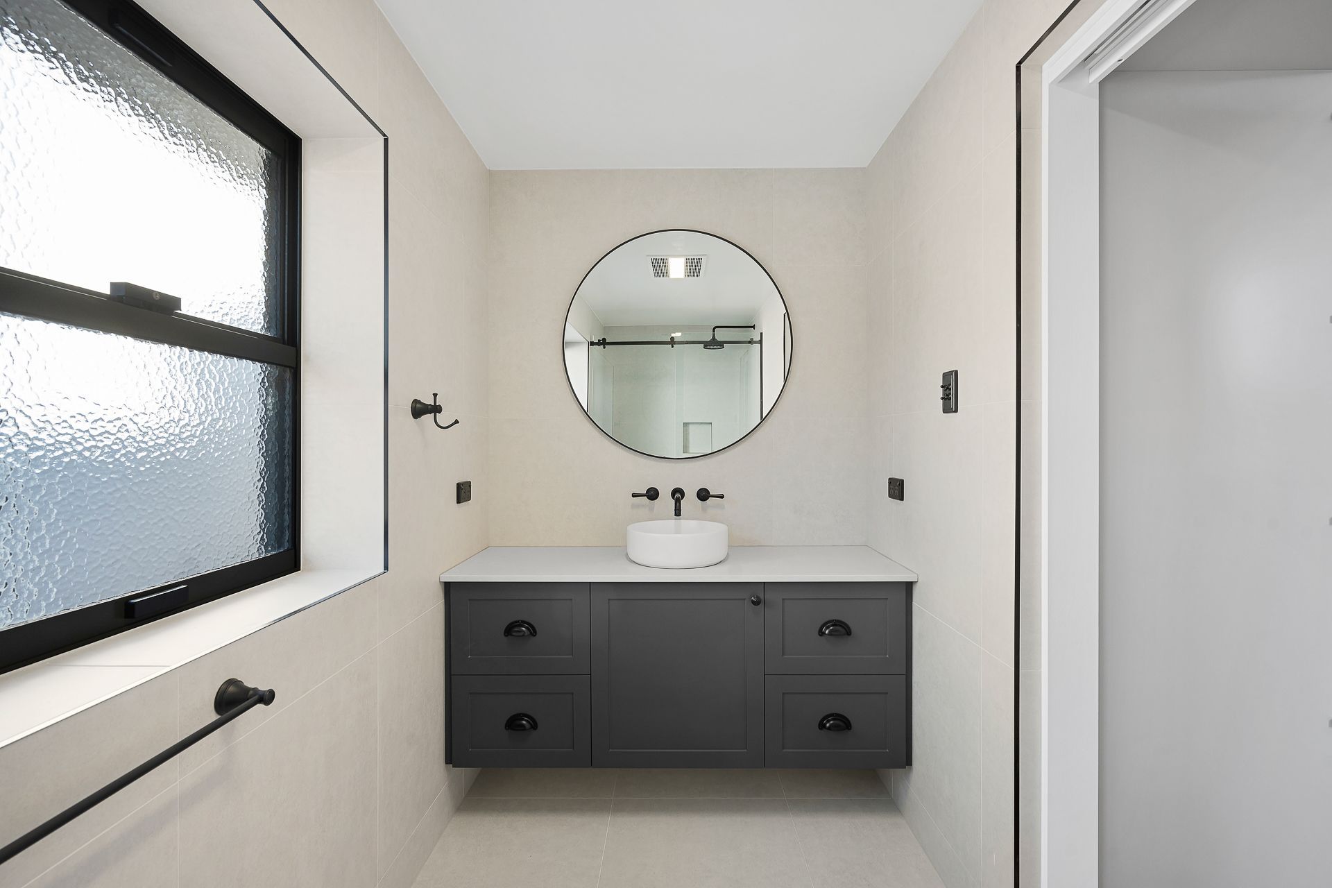A modern bathroom with a charcoal floating vanity, a round mirror, a white vessel sink, and a black-framed window.
