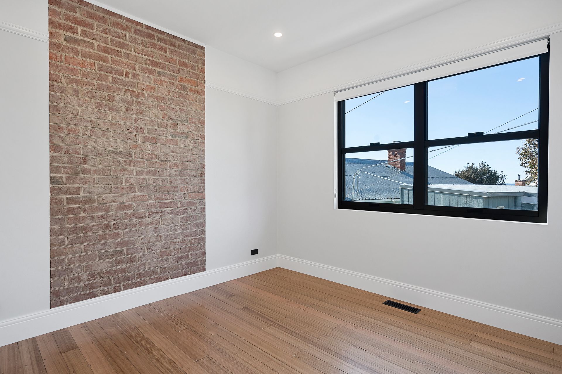 An empty room with light wood floors, grey walls, a vertical exposed brick accent wall, and a large black-framed window.