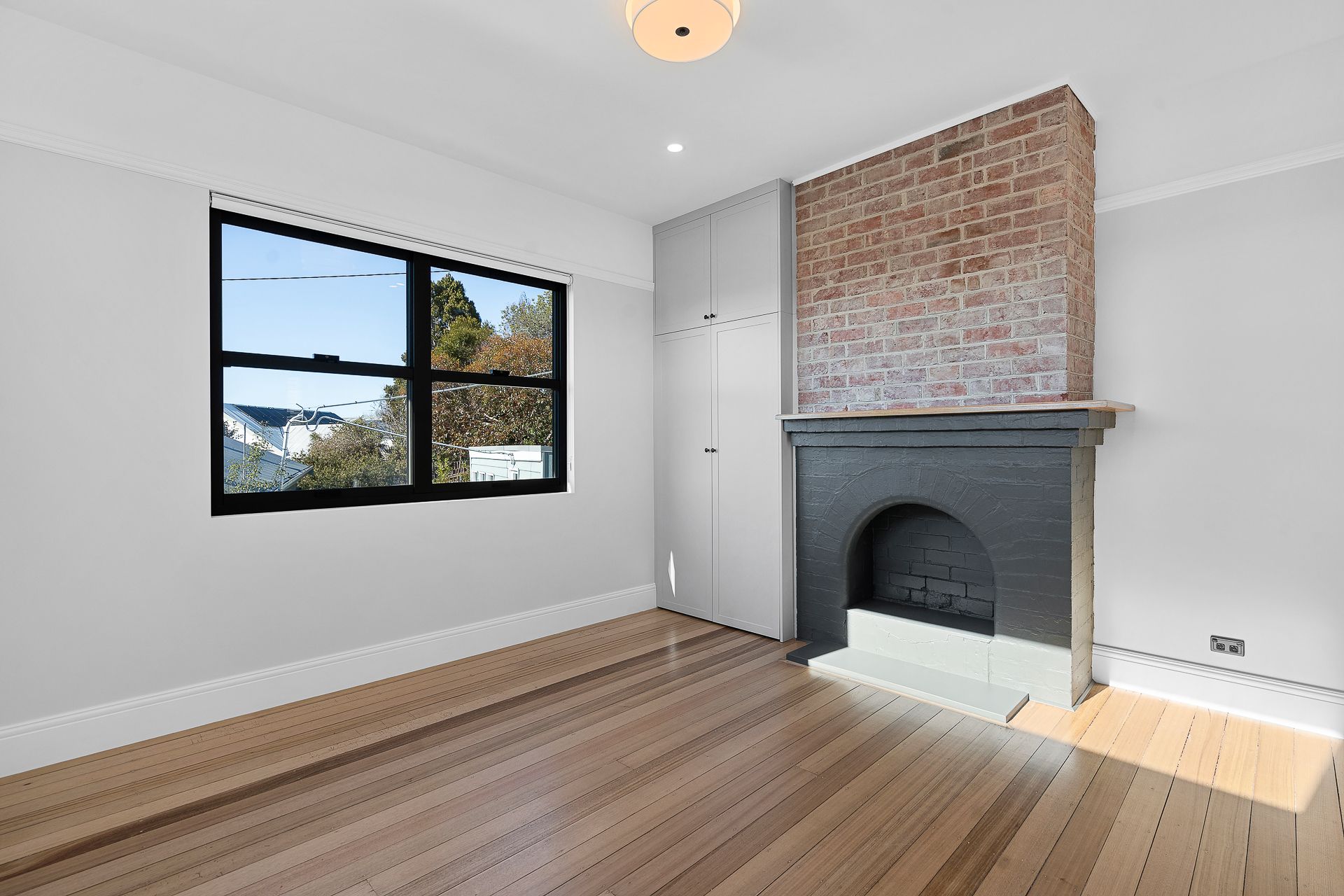 Empty room with light wood floors, gray walls, a black window, and a fireplace with exposed brick and a black base.