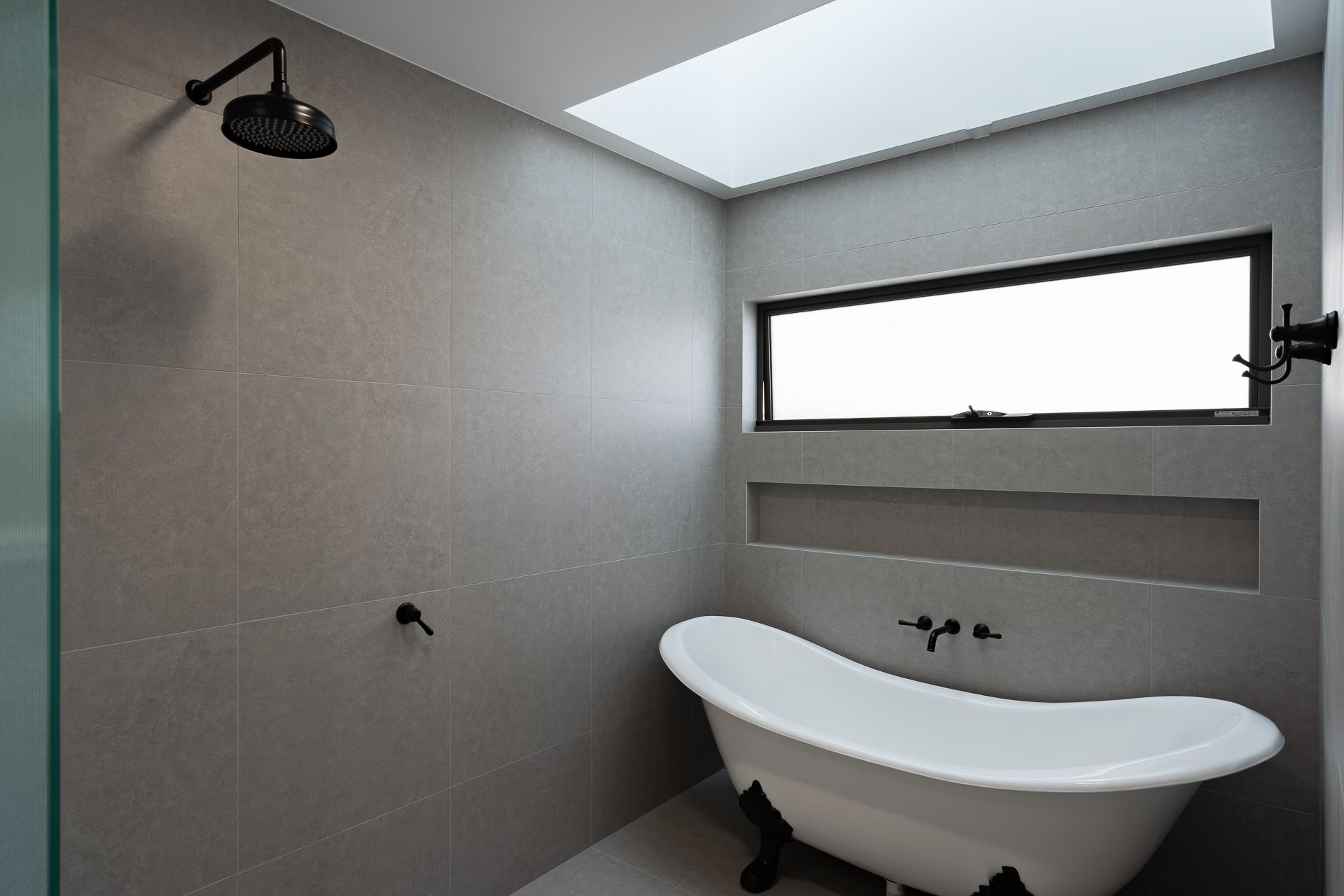 A modern bathroom with grey tiled walls, a white claw-foot tub, a rainfall showerhead, and a rectangular window.