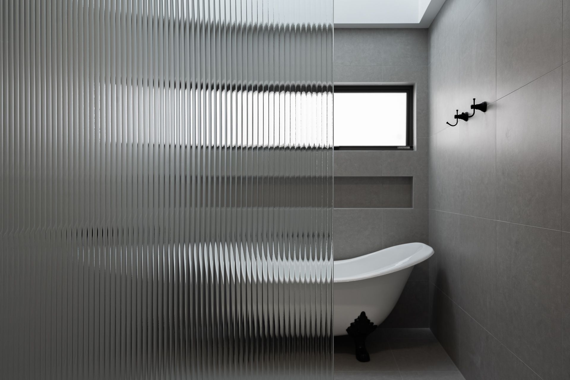 A white claw-foot bathtub sits in a modern gray bathroom behind a textured, vertical-lined glass partition.