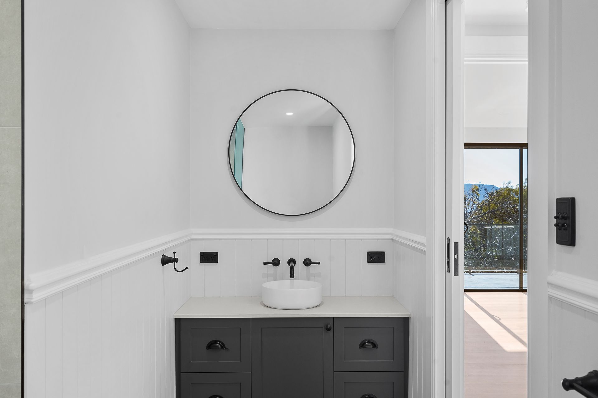 A modern powder room featuring a dark vanity with a white basin, a round mirror, and white wainscoting on the walls.