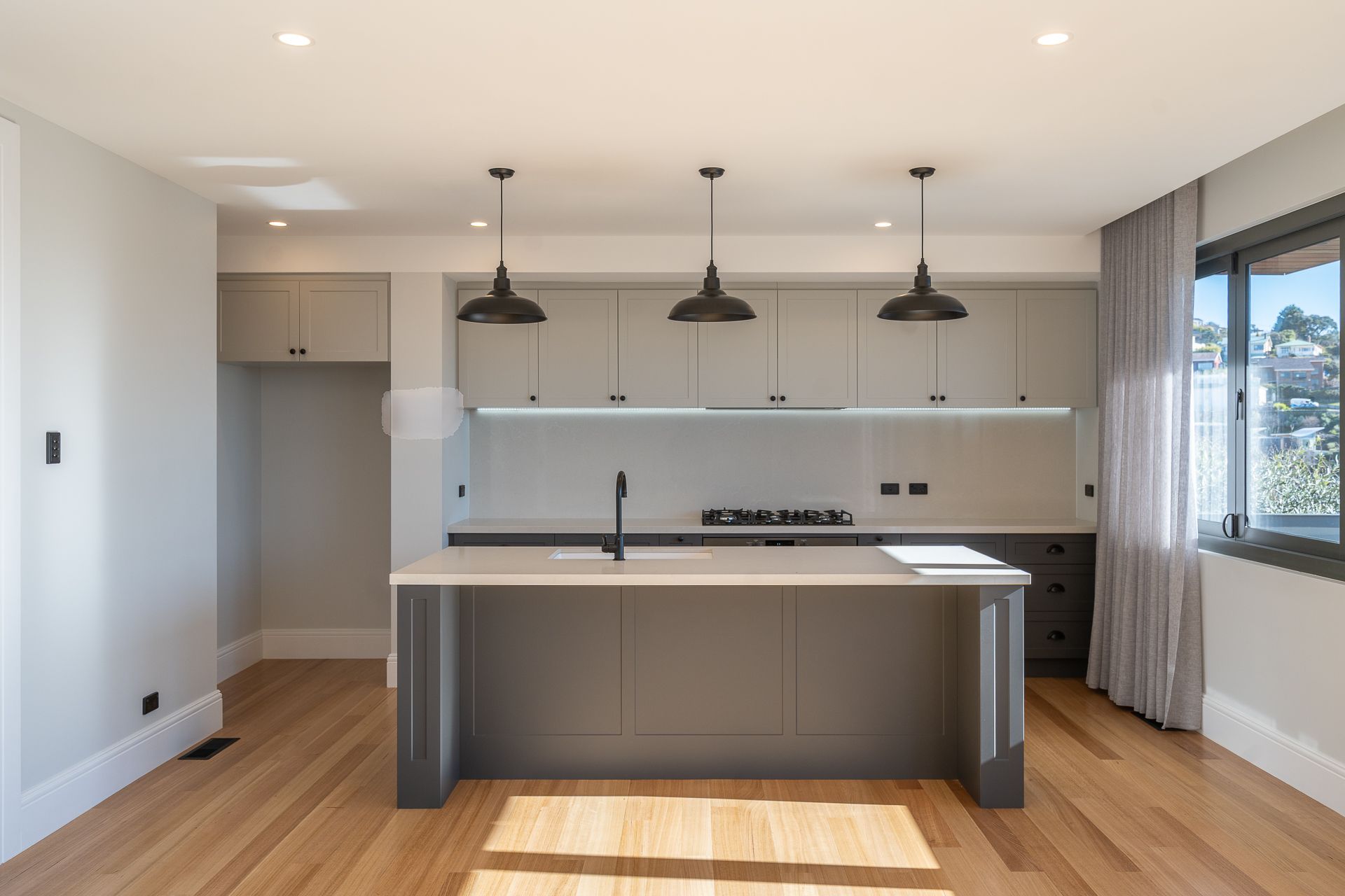 Modern kitchen with a dark grey central island, light grey cabinets, black pendant lights, and light wood flooring.
