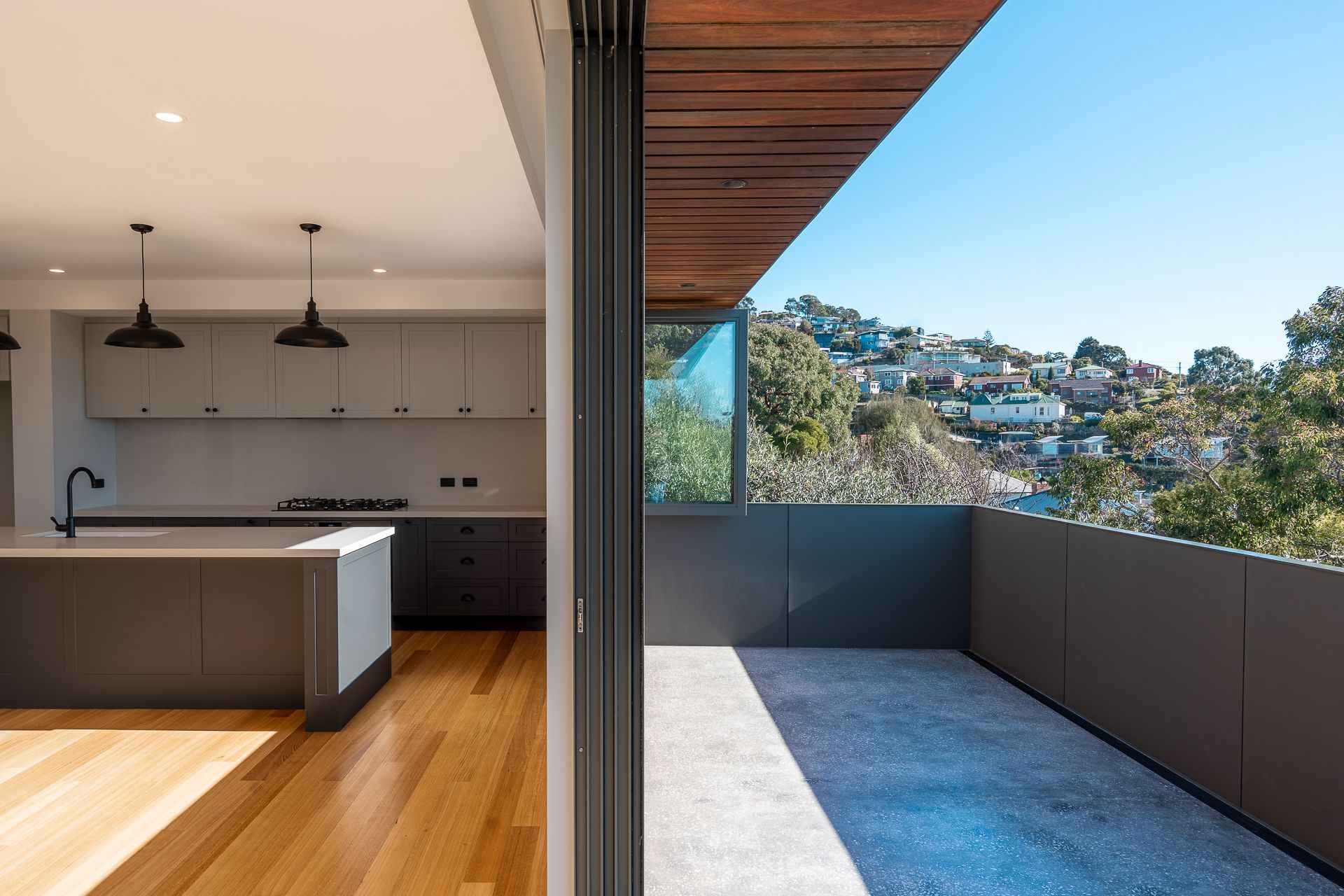 Modern kitchen with hardwood floors opening to a balcony overlooking a hillside neighborhood under a clear blue sky.