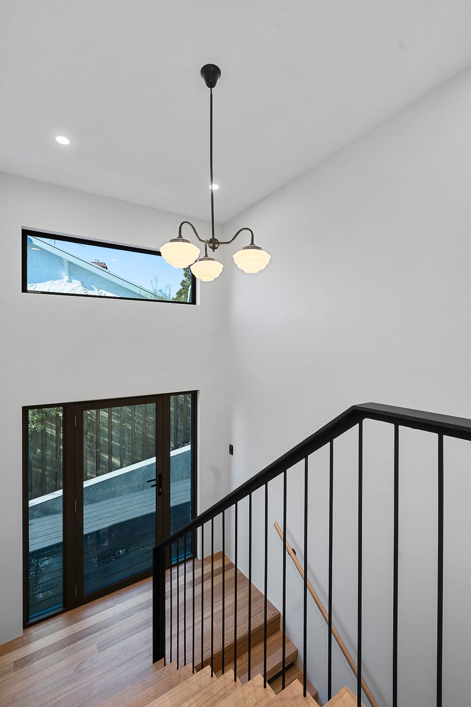 A modern entryway with a light wood staircase, black metal railing, a hanging three-globe chandelier, and tall windows.