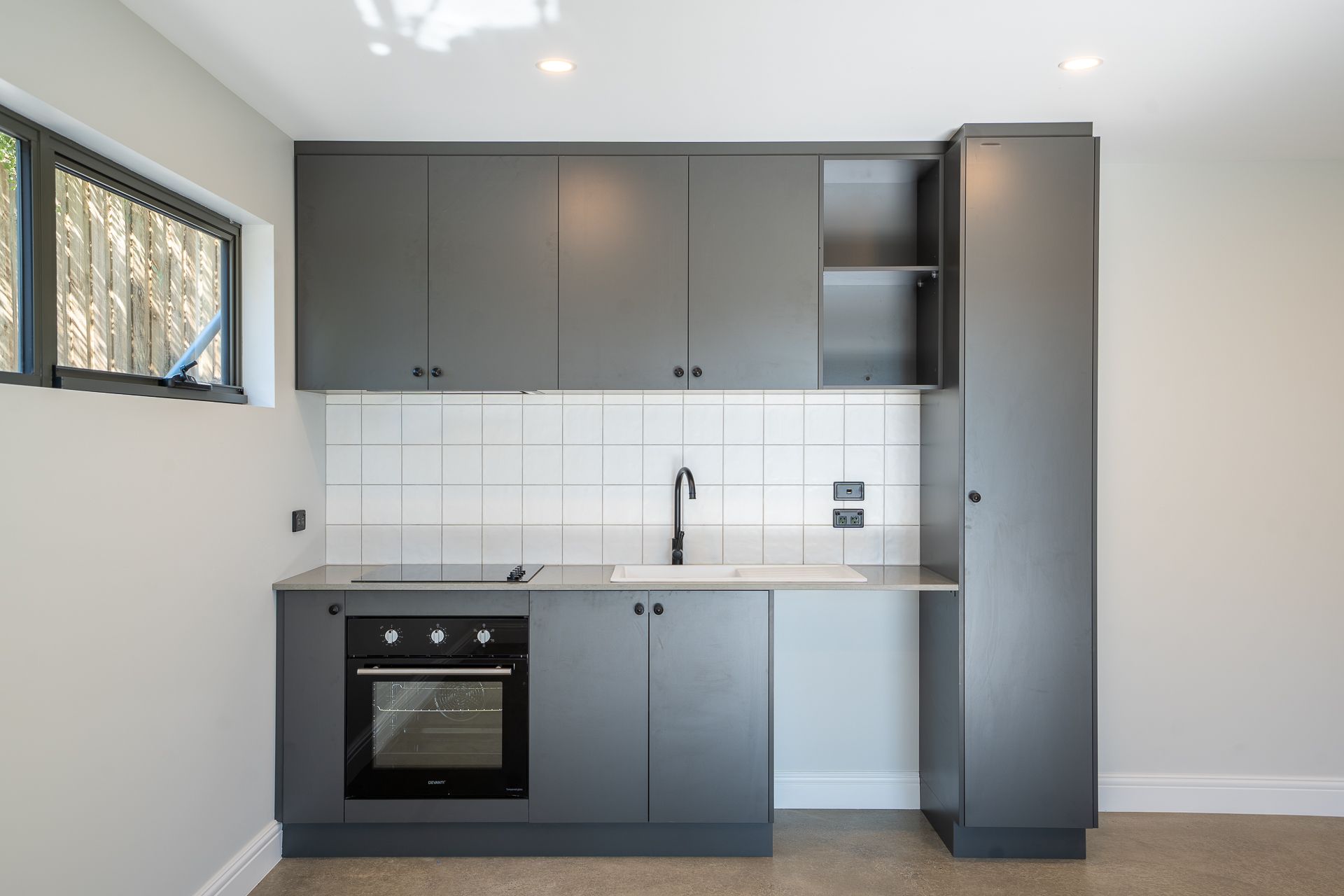 A modern kitchen featuring grey cabinets, white tiled backsplash, built-in oven, sink, and a tall pantry cupboard.