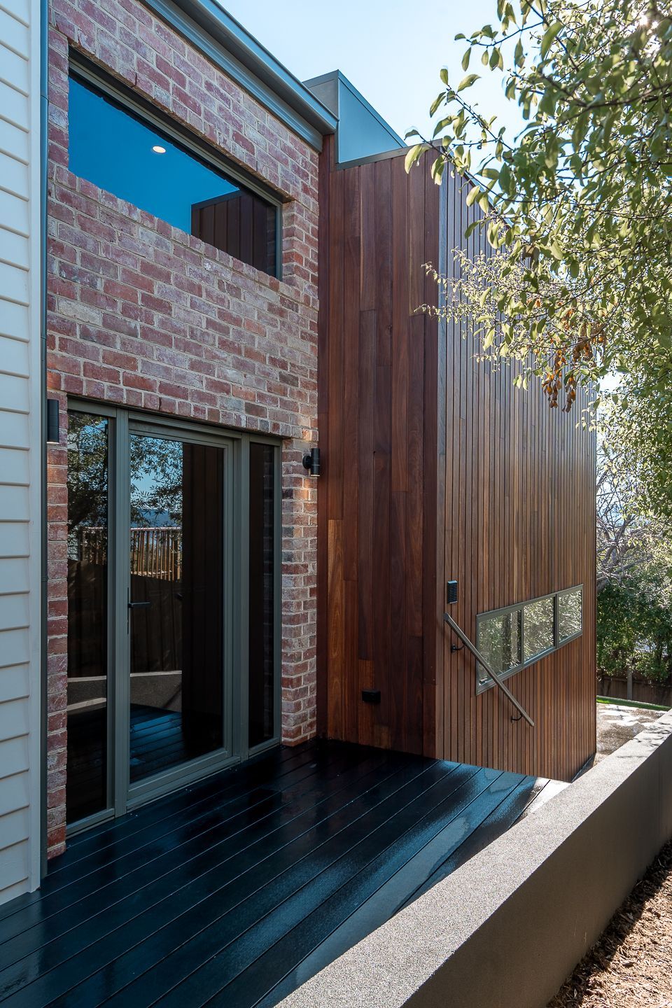 A multi-story house exterior featuring a red brick facade, a wooden panel wall, a black deck, and sliding glass doors.