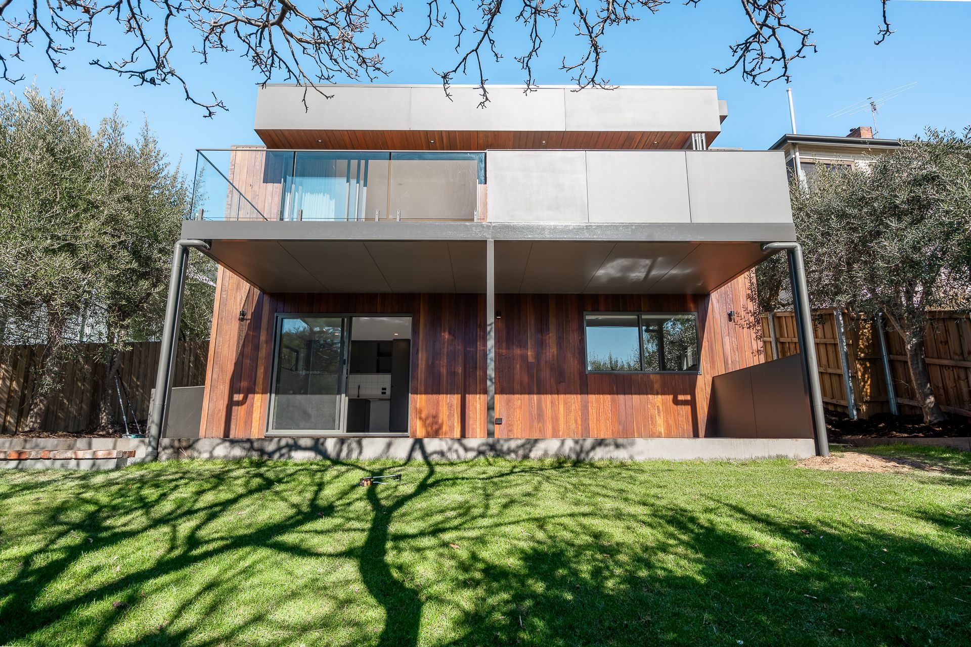 Modern two-story house with wood siding, a balcony, and sliding glass doors, viewed from a sunny backyard lawn.
