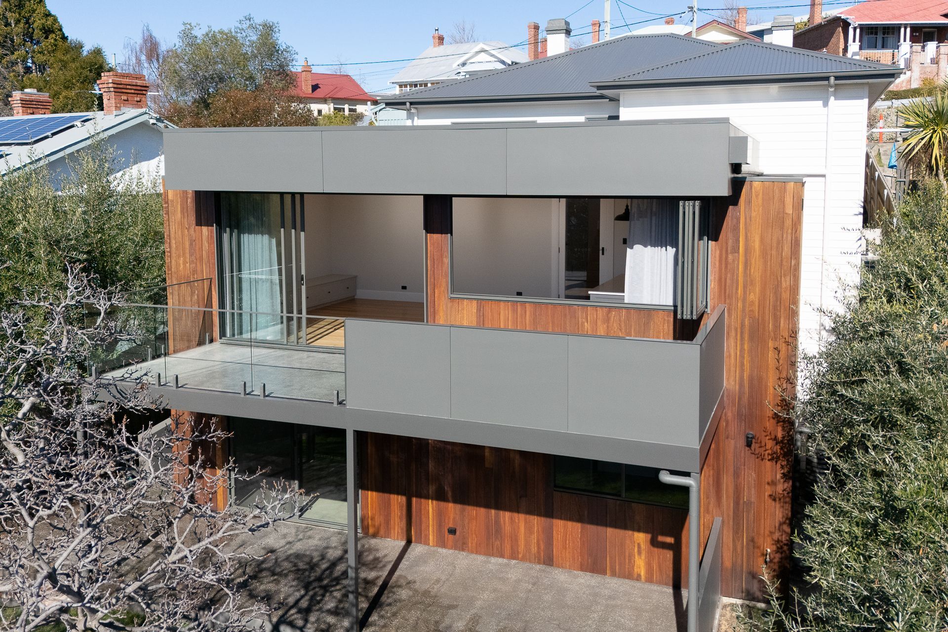 A contemporary, two-story house extension with wood siding and a flat grey roof, featuring an open balcony and glass doors.